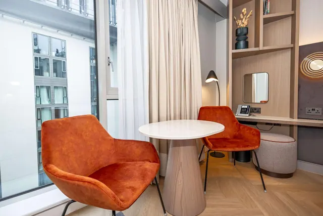 Two orange velvet armchairs sit beside a round white table in front of a large window overlooking a neighbouring building, with a desk and mirror to the right.