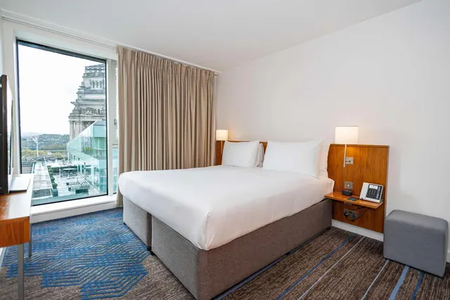 A made-up double bed with white linen faces a large window showing a city building and rooftops outside.