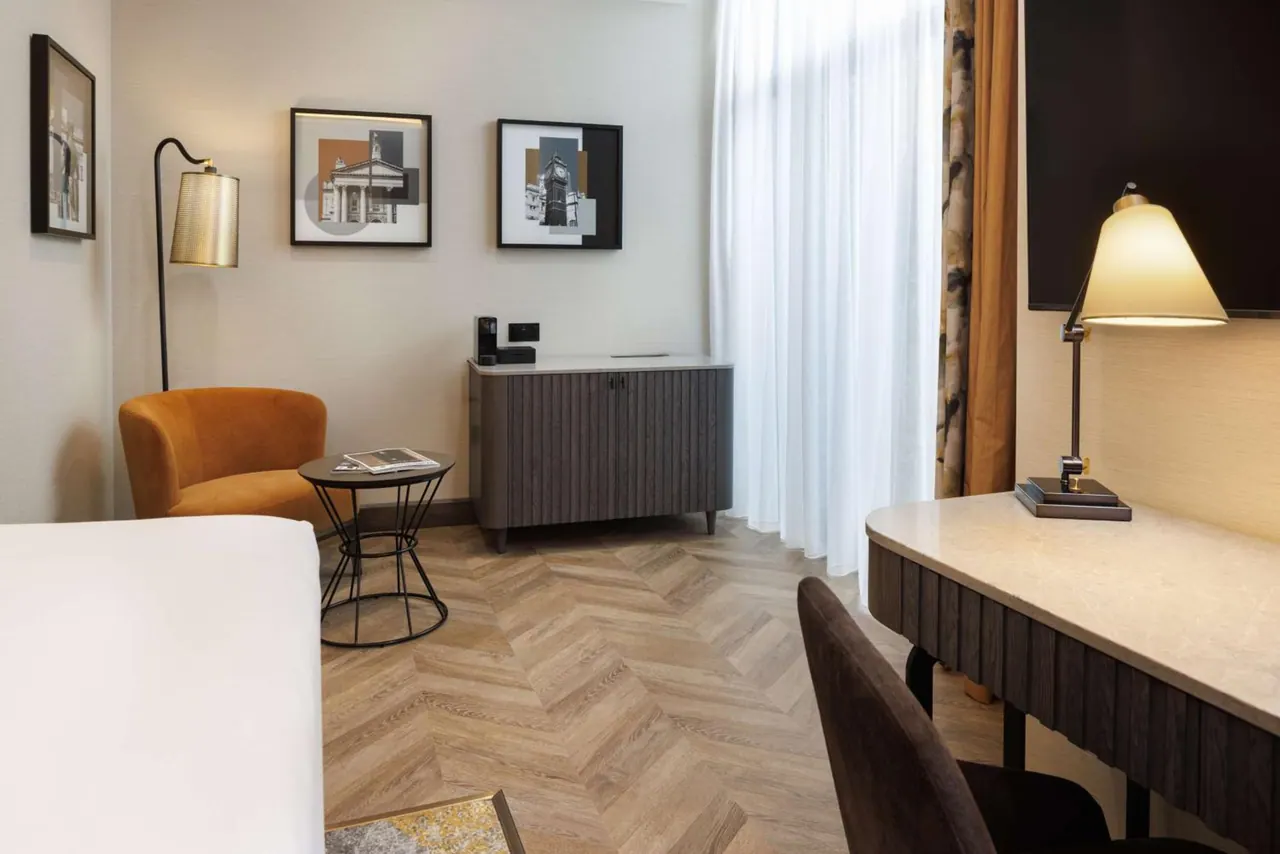 A hotel bedroom with an orange armchair and round side table, a wood-fronted sideboard beneath framed prints, a desk with a table lamp, and a wall-mounted television.