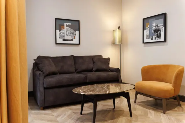 A living area with a dark brown sofa, an orange armchair, a marble-topped coffee table, a floor lamp and two framed prints on the walls.