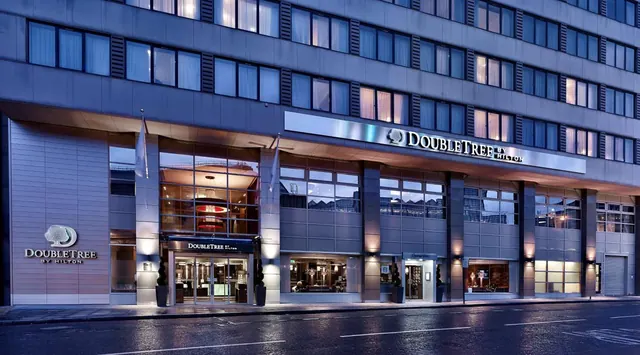 The exterior of a DoubleTree by Hilton hotel at dusk, with lit windows, a glass entrance and a wet road in front.