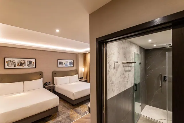 A hotel bedroom with two double beds and framed wall art, with an open doorway to a bathroom containing a walk-in glass shower with marble-look walls.