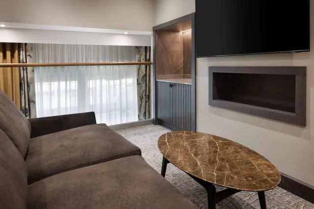A hotel living area with a brown sofa, an oval marble-topped coffee table, a wall-mounted television and a window with sheer curtains.
