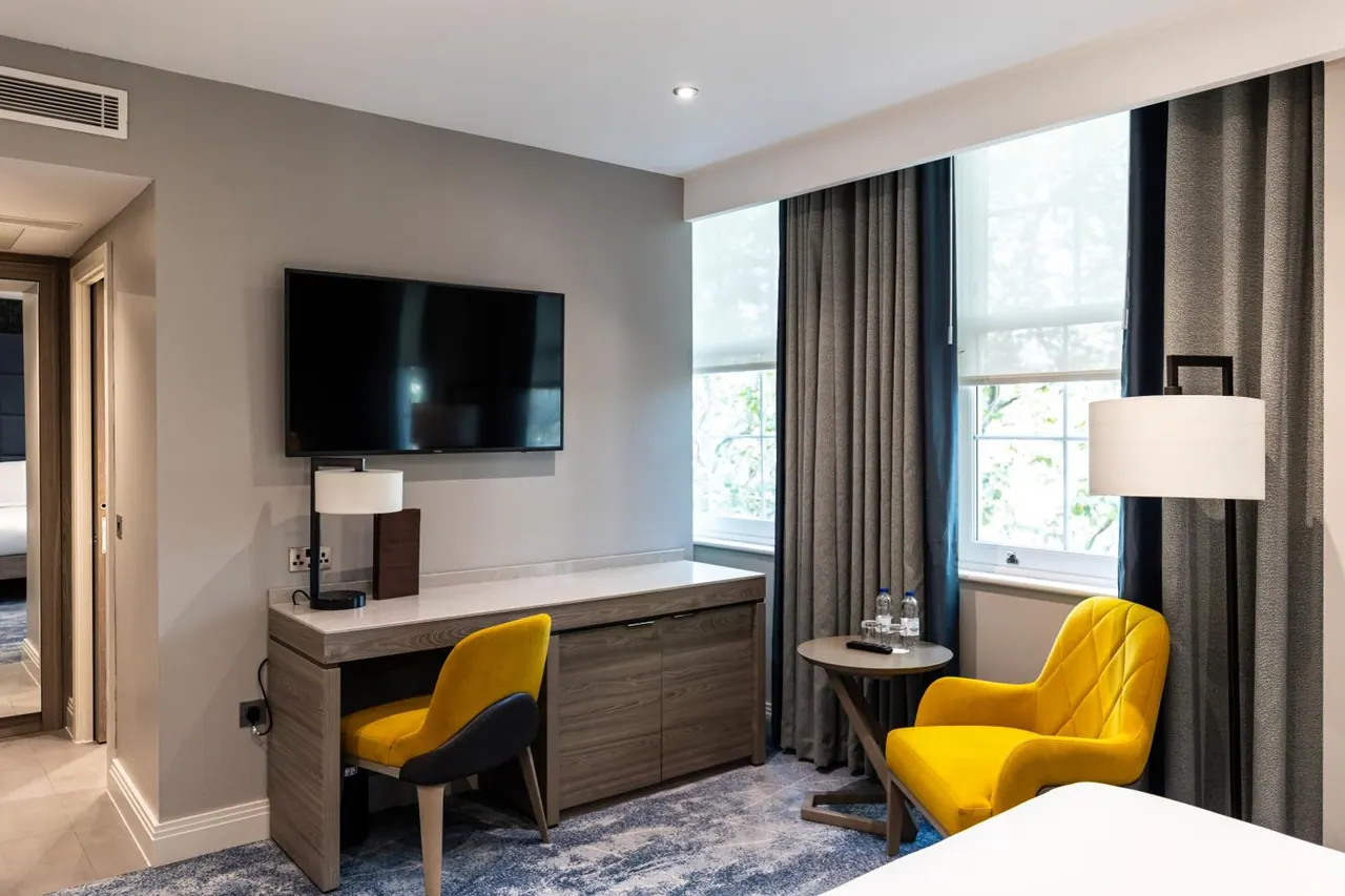 A hotel bedroom with a wall-mounted television, a work desk, two yellow armchairs and a side table with bottled water beside tall windows.