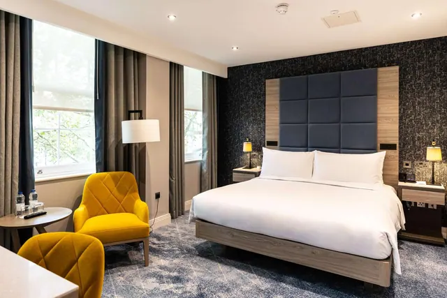 A bedroom with a large white-covered double bed, a dark tufted headboard, two yellow armchairs by the window and a small round table with water bottles.