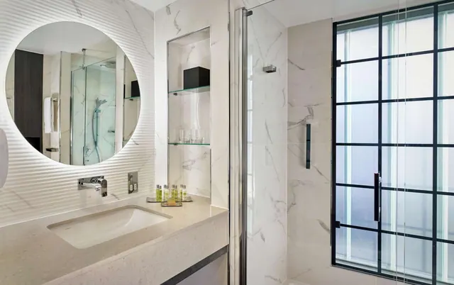 A bathroom with a round backlit mirror, a single undermount sink, a glass-enclosed walk-in shower and a tall black-framed frosted window.