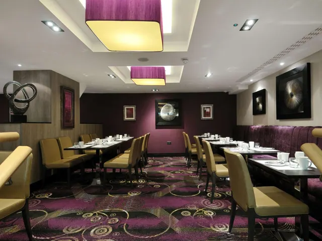 A restaurant dining room with rows of set tables, tan leather chairs, a purple upholstered banquette and a patterned purple carpet.