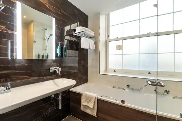 A bathroom with a wall-mounted basin, a lit mirror, dark tiled walls, and a bathtub behind a glass screen beside a large frosted window.