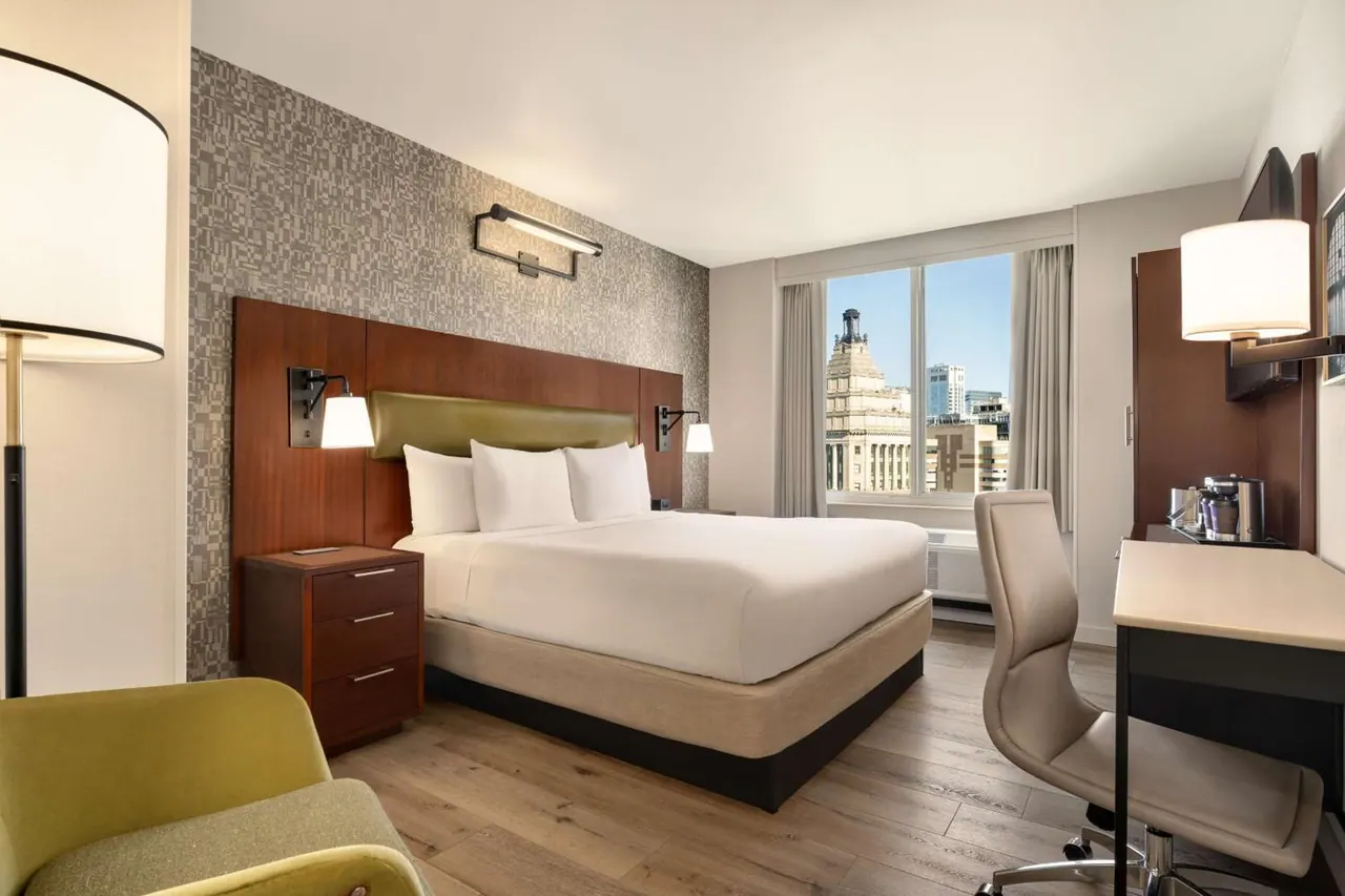 A hotel bedroom with a king bed, a green armchair, a work desk and a window looking out over city buildings.
