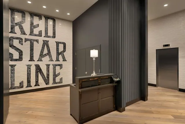A lobby with large Red Star Line lettering on a white brick wall, a reception desk with a table lamp and card terminal, and a lift on the right.