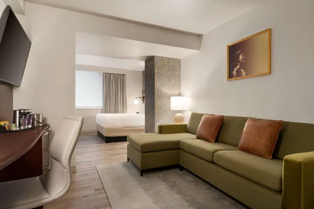 A living area with a green sectional sofa, brown cushions, a table lamp and framed artwork, with a desk holding a coffee station on the left and a bed visible through the doorway.