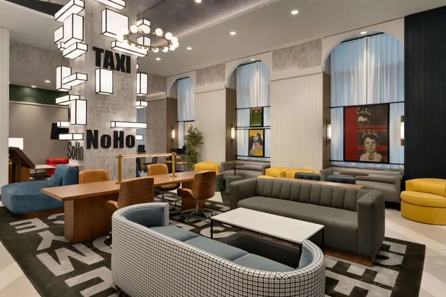 A lobby lounge with grey and yellow sofas, armchairs, a long wooden table, and TAXI and NoHo signs on a pillar.
