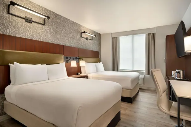 A hotel room with two queen beds, a wall-mounted television, a window with a blind, and a desk with a chair.