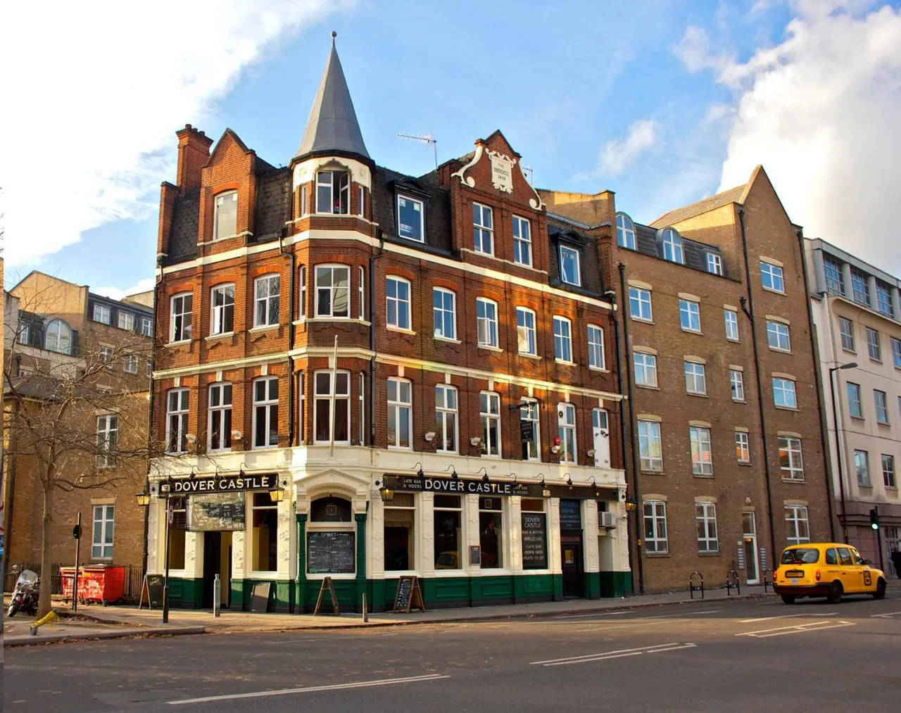 The exterior of a brick Victorian corner building with a spire and a Dover Castle pub frontage, with a yellow taxi on the street beside it.