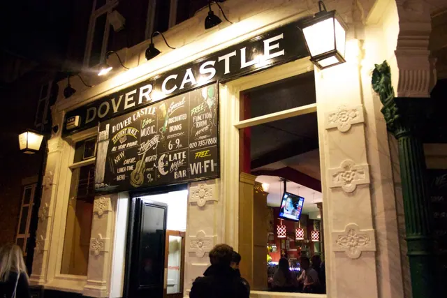 Night-time exterior of the Dover Castle pub with its illuminated fascia sign, chalkboard notices and open doorway, with a few people visible in the foreground and inside.