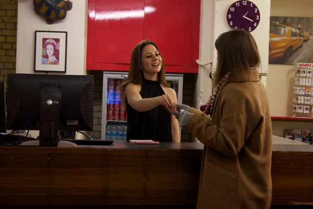 A woman behind a reception desk shakes hands with a guest, with a computer monitor, a drinks fridge and a red wall cabinet behind the desk.