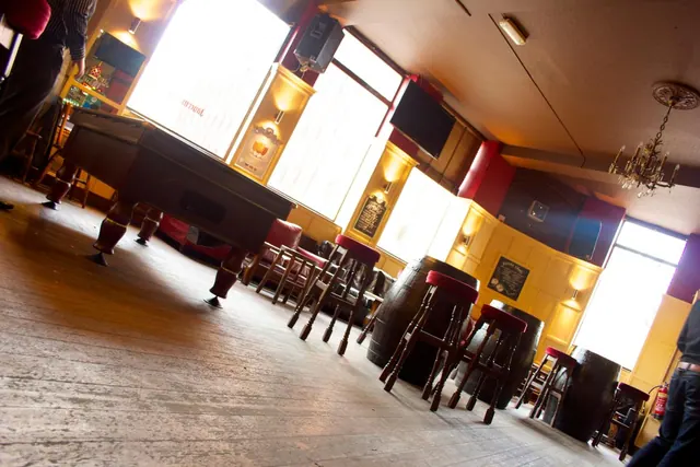 A pub-style room with a pool table, red-topped bar stools, barrel tables, a wall-mounted television and bright windows, photographed at a tilt.