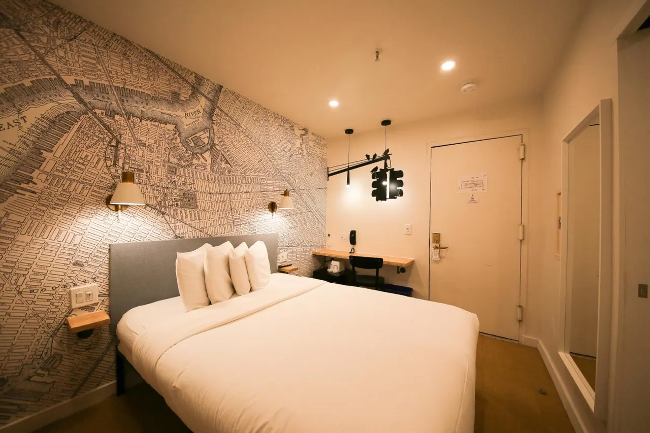 A bedroom with a map-printed feature wall, a double bed with white bedding, a small wall-mounted desk with a chair, and a closed door.