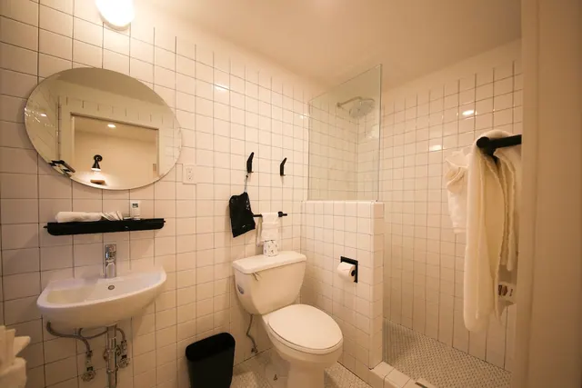 A bathroom with white tiled walls, a round mirror above a wall-mounted sink, a toilet, and a glass-enclosed walk-in shower with folded white towels.
