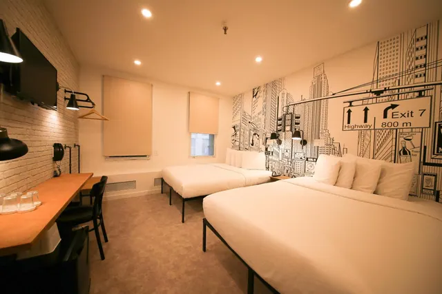 A hotel bedroom with two double beds, a black and white city skyline mural with an Exit 7 highway sign on the wall, a wall-mounted television and a wooden work desk.