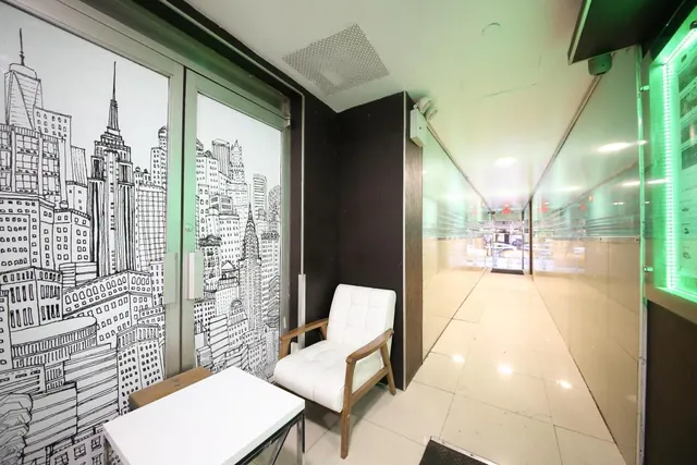 A small entrance area with a white armchair and white table beside a window mural of a city skyline, with a glass-walled corridor leading toward the street.