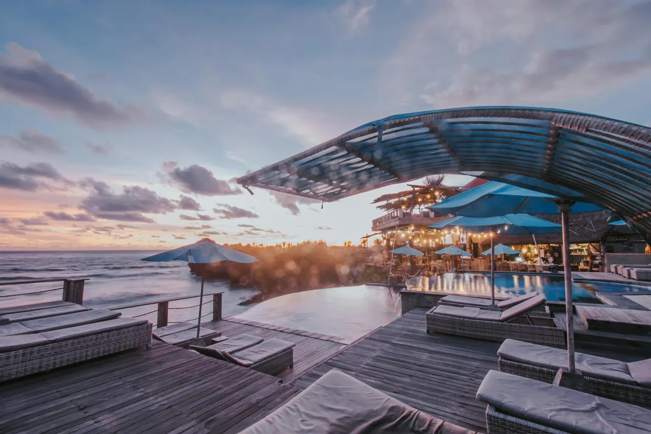 An infinity pool and wooden deck with sun loungers and blue umbrellas overlook the ocean at sunset.