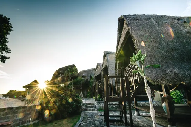 A row of thatched-roof bungalows with wooden stairs and decks stands beside a stone path, with the low sun flaring through the trees at left.