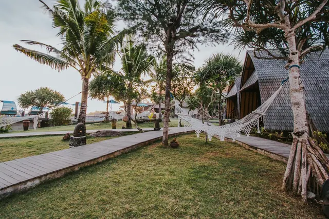 A wooden walkway runs across a grassy garden lined with palm trees, with a white hammock strung between trees and shingle-roofed bungalows on the right.