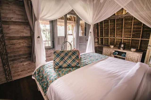 A bedroom with a white canopy mosquito net over a bed dressed in white linen and a teal batik runner, with open doors onto trees and a bamboo-clad wall.