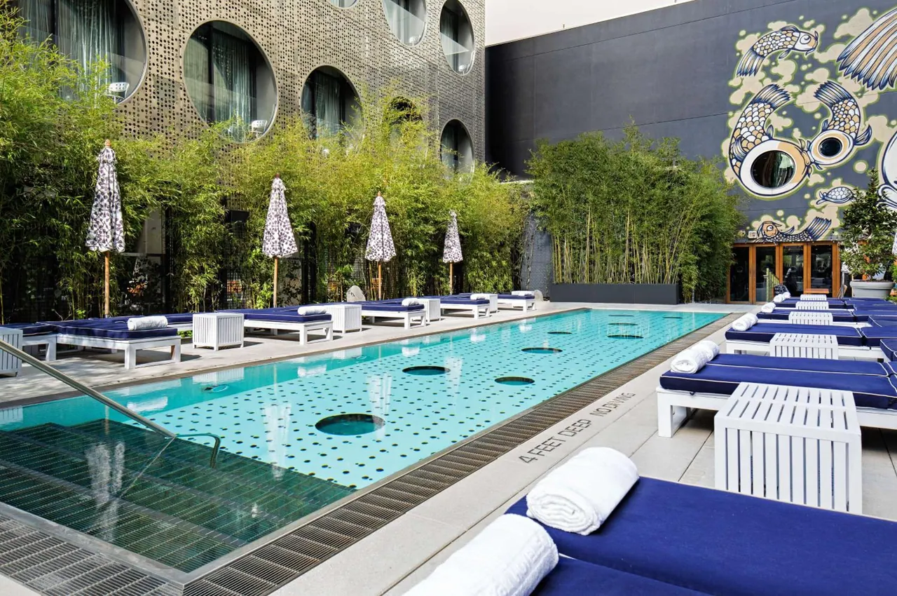 An outdoor pool with blue-cushioned loungers, white towels, folding umbrellas, bamboo planting and a koi mural on the wall behind.