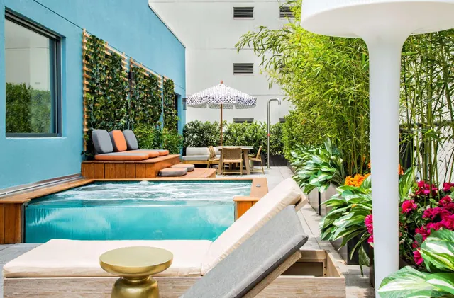 A small blue pool with bubbling jets sits on a wooden deck beside a vertical plant wall, a cushioned lounger, and a striped umbrella.