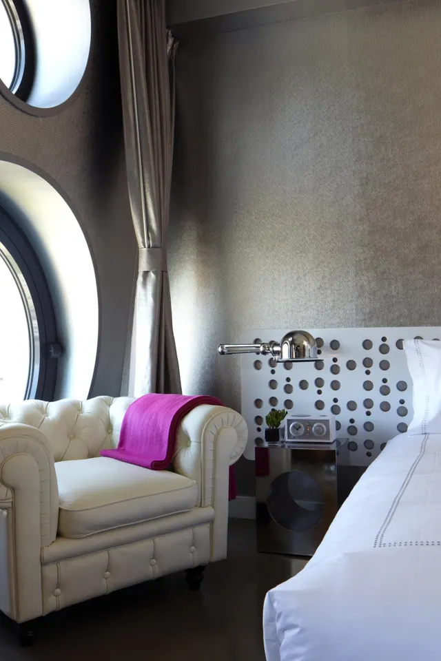 A bedroom with round porthole windows, a cream tufted armchair with a magenta throw, a perforated headboard and a white bed.