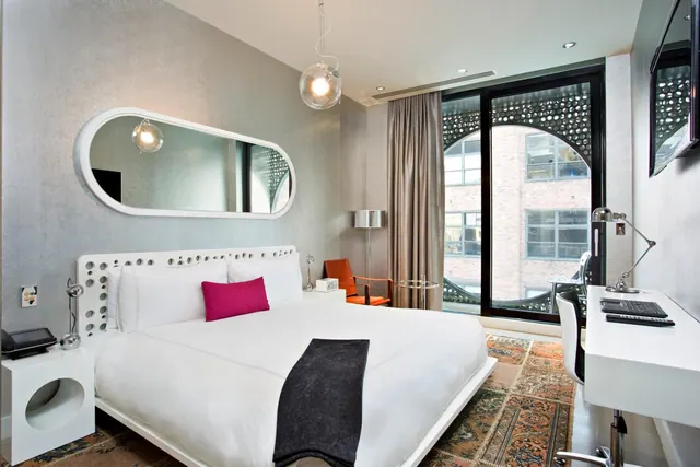 A bedroom with a king bed, a pink cushion, a white desk, an orange chair and floor-to-ceiling windows looking onto a brick building.