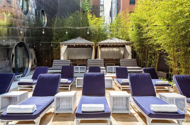 Rows of navy sun loungers with folded white towels and white side tables sit on a sand-coloured outdoor area beside bamboo and two canopied cabanas, under strung lights.