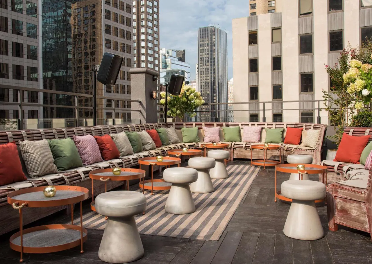 A rooftop terrace with cushioned banquette seating, concrete stools and low orange tables, with tall city buildings beyond the railing.