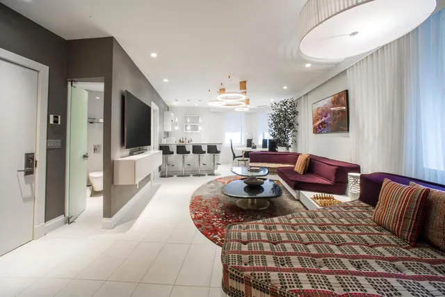 A modern living area with a purple sofa, a patterned sectional sofa, a wall-mounted television, a round coffee table, and a bar counter with stools in the background.