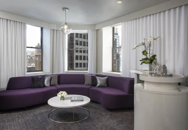 A living area with a curved purple sofa, a round white coffee table, and windows looking out onto city buildings.