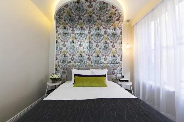 A bedroom with a double bed in front of an arched patterned wallpaper wall, with a white side table on each side and sheer curtains at the window.