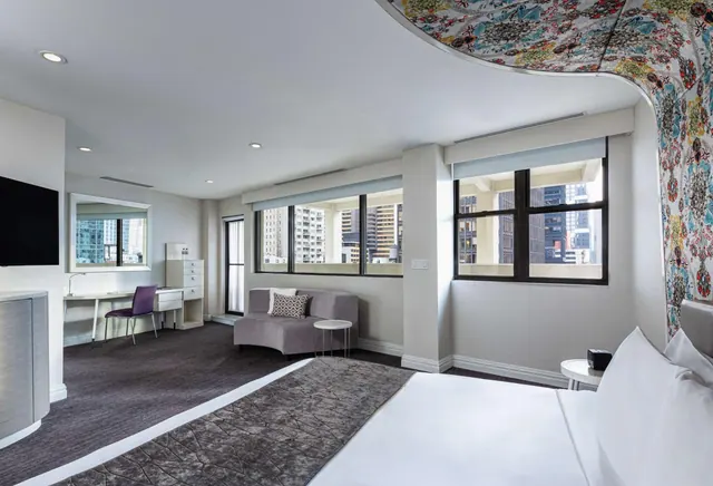 A hotel bedroom with a made bed in the foreground, a grey curved sofa, a desk with a purple chair, and windows looking onto city high-rises.