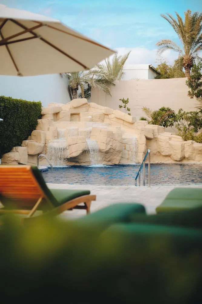 A pool with a rock-style waterfall feature at the far side, with a parasol and green sun loungers in the foreground and palm trees behind.