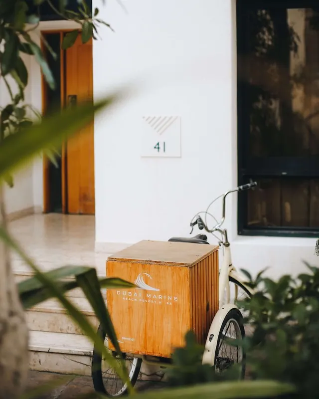 A white building entrance with a numbered door sign and a wooden cargo tricycle branded Dubai Marine Beach Resort & Spa parked outside, with foliage in the foreground.