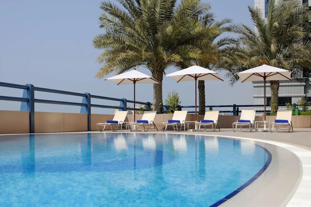 An outdoor pool with a curved edge, white umbrellas and blue-cushioned sun loungers, with palm trees and a building behind.