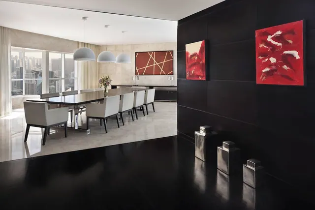 A dining area with a long dark table, grey upholstered chairs, white pendant lights, red wall paintings, and large windows showing high-rise buildings outside.