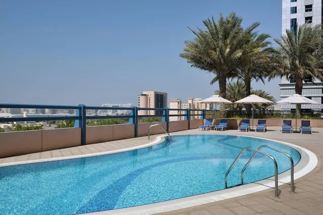 A rooftop pool with blue loungers and white umbrellas, with palm trees and city buildings behind it under a clear sky.