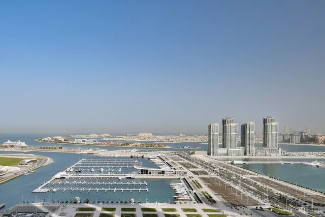 Aerial view over a marina with moored boats, white residential towers and a distant island under a clear blue sky.