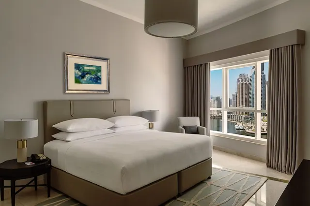 A made king bed sits in a bedroom with a framed painting, an armchair, and a window overlooking high-rise towers and a marina.