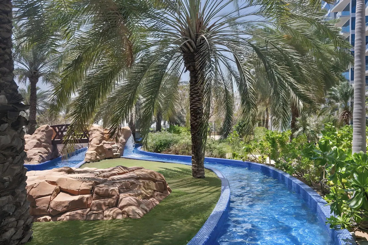A winding blue lazy river runs past palm trees, a rock formation and a green lawn, with a high-rise building at the right edge.