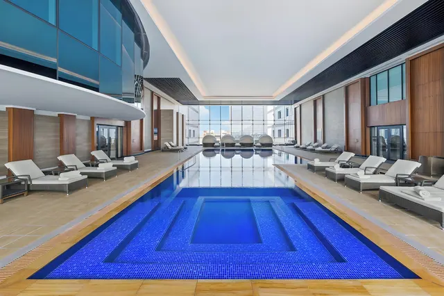 Indoor pool with blue tiled steps, white-cushioned loungers along both sides, and floor-to-ceiling windows showing buildings at the far end.