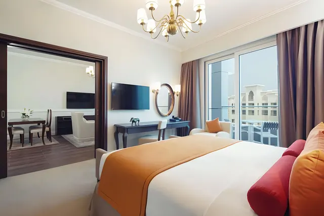 A bedroom with a king bed under an orange throw, a desk with a round mirror, a wall-mounted television, and a sliding door opening onto a balcony with buildings beyond.
