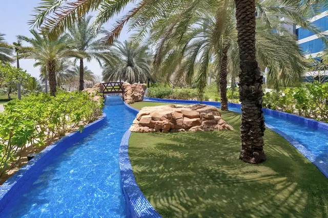 A blue-tiled water channel runs between palm trees, a rock feature on a grass lawn, and a wooden footbridge in the background.